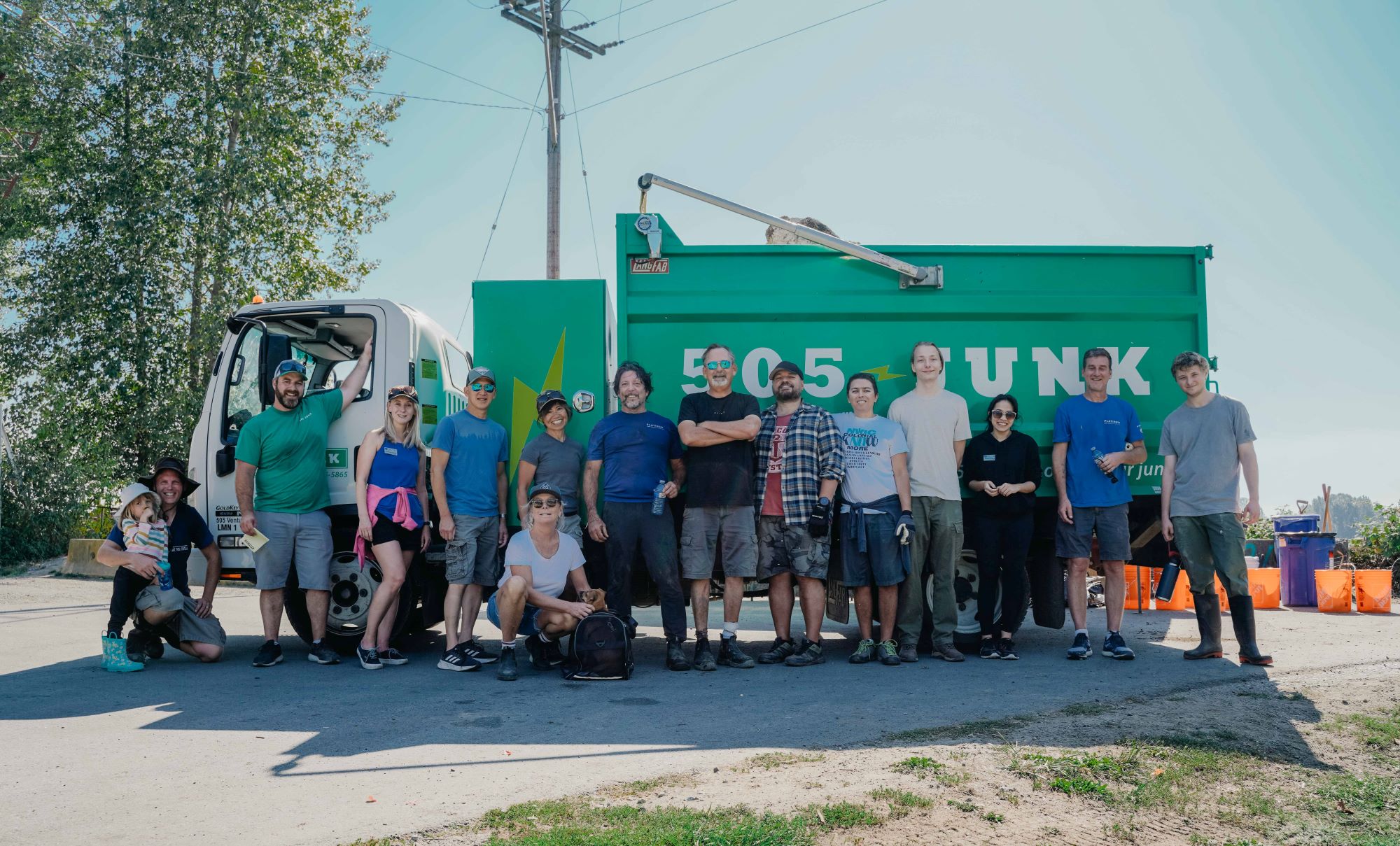 A group of volunteers from Platinum Pro-Claim Restoration and 505-Junk standing in front of their trucks during a community cleanup event, holding orange buckets and wearing gloves, with a clear blue sky in the background.